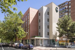 A multi-story hotel with a prominent clock tower, a parking lot with several cars, and a street with trees. - B&B Hotel Zaragoza Plaza Mozart (Zaragoza)
