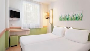 A hotel room with a bed, a TV mounted on the wall, a desk with a chair, and a window with curtains.