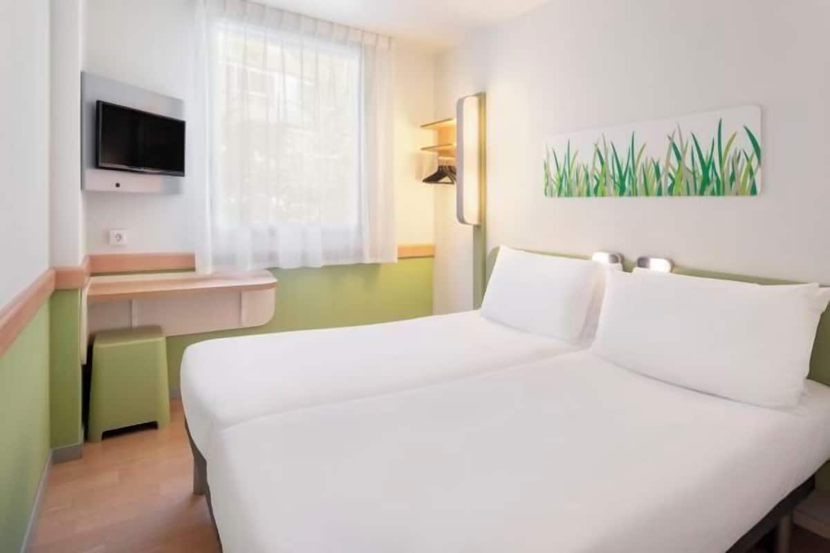 Ein Hotelzimmer mit Bett, Wandfernseher, Schreibtisch mit Stuhl und Fenster mit Vorhängen.