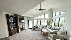A spacious living room with a fireplace, large windows, and a ceiling fan. - Brooklyn Bridge Vacation Condo (Arnolds Park)