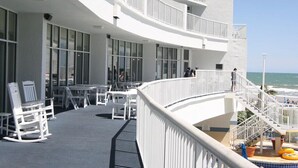 A curved deck with white railings, outdoor seating, and a pool area with lounge chairs and umbrellas. - Sea Watch Resort (Myrtle Beach)