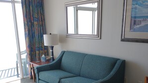 A hotel room with a teal sofa, a wooden coffee table, a round dining table, a lamp, and a framed picture on the wall. - Sea Watch Resort (Myrtle Beach)