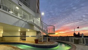 A swimming pool area with a curved design, a staircase leading to an upper level, and a sunset view. - Sea Watch Resort (Myrtle Beach)
