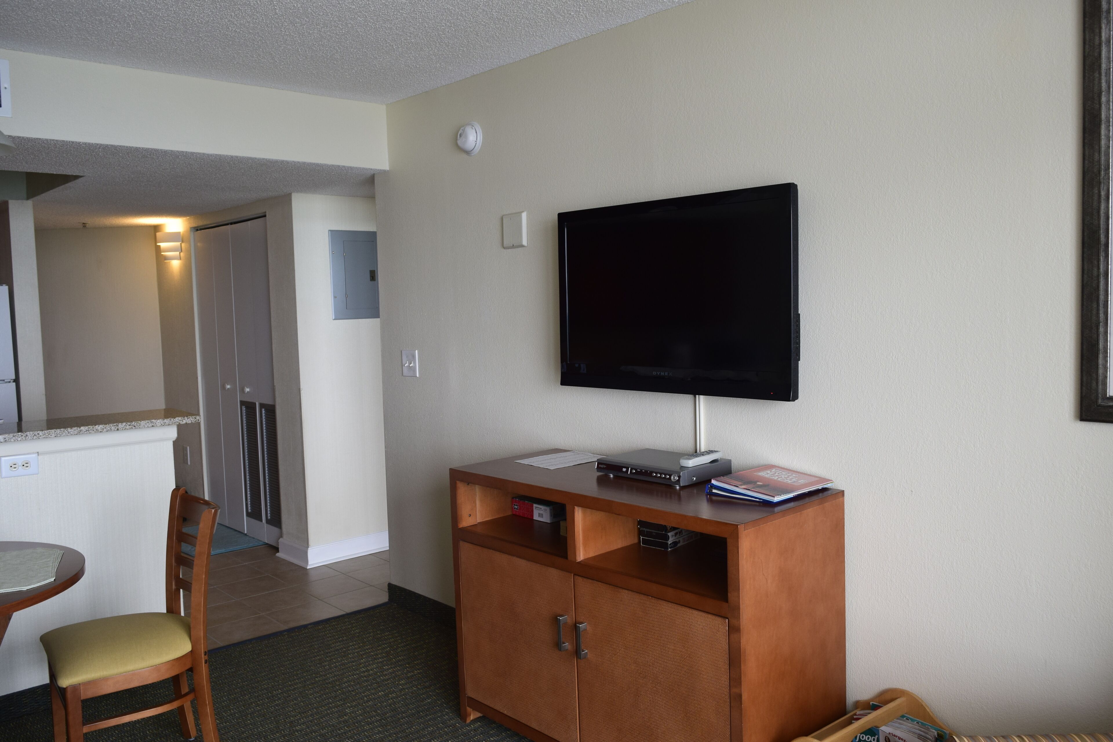 A hotel room with a kitchenette, a flat-screen TV mounted on the wall, a wooden cabinet, and a dining area with a chair and table.