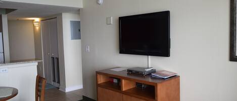 A hotel room with a kitchenette, a flat-screen TV mounted on the wall, a wooden cabinet, and a dining area with a chair and table.