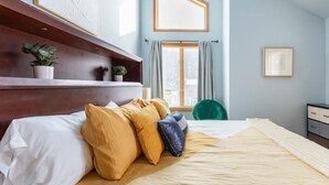 Une chambre à coucher avec un lit, des oreillers, une table de chevet, une commode, une fenêtre avec des rideaux et une plante.