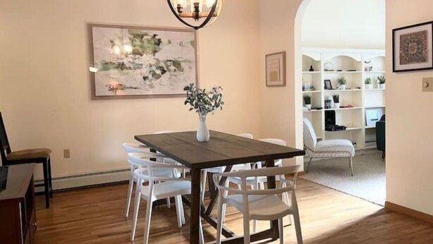 Un coin repas avec une table en bois, des chaises blanches, un vase de fleurs et un luminaire suspendu.