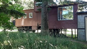 A wooden cabin with a small attached shed, surrounded by tall grass and trees.