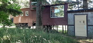 A wooden cabin with a small attached shed, surrounded by tall grass and trees.