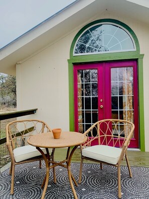 Een terras met twee rieten stoelen en een kleine tafel, een kleurrijke deur met een groene omlijsting, en een vloer met een patroon.