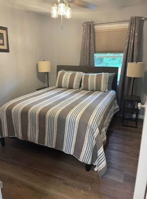 A bedroom with a bed, two bedside lamps, a window with curtains, a ceiling fan, and a door.