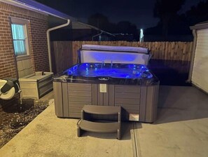 A hot tub with a built-in seat and a step for easy access, located in a backyard with a wooden fence and a brick house in the background.