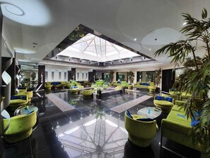 Spaziosa hall con soffitto alto e lucernario, caratterizzata da aree relax con sedute verdi e piante in vaso.