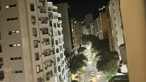 A nighttime view of a city street lined with high-rise buildings, featuring a well-lit road with vehicles and trees. - Ap Copacabana 200m da praia – CP4 (Rio de Janeiro)
