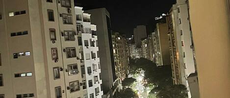 Une vue nocturne d’une rue de la ville, bordée d’immeubles de grande hauteur, avec une chaussée bien éclairée, des véhicules et des arbres.