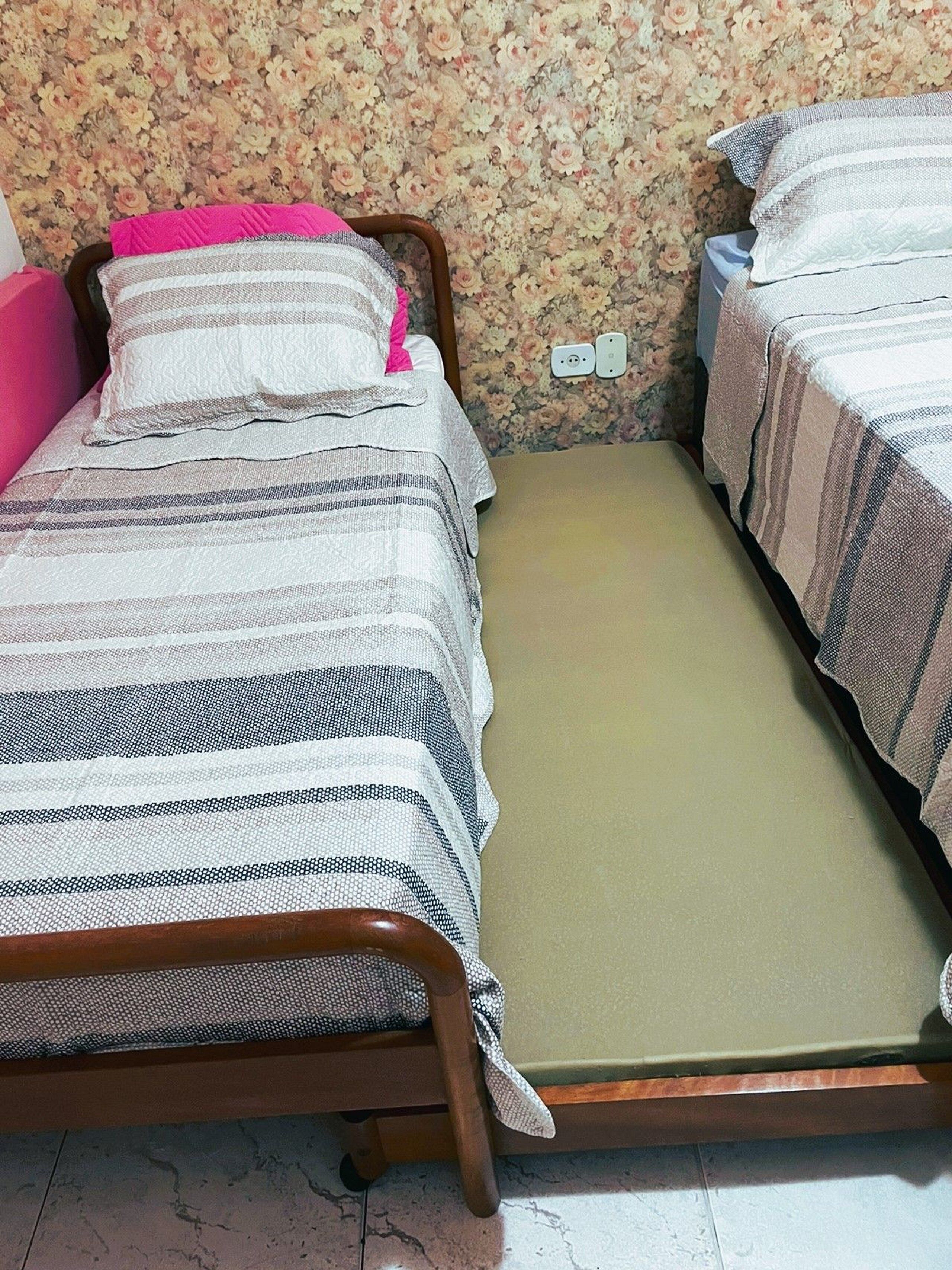 A room with two beds, one with a wooden frame and the other with a metal frame. There is a floral wallpaper on the wall and a power outlet visible.