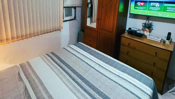 A bedroom with a bed, a wooden wardrobe, a television displaying a sports channel, and a window with blinds. - Ap Copacabana 200m da praia – CP4 (Rio de Janeiro)