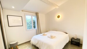 Een slaapkamer met een bed, een klein krukje, een nachtkastje, een lijst met een afbeelding, een raam met gordijnen en een wandlamp.