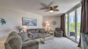 Ein Wohnzimmer mit grauem Sofa, Couchtisch, Lampe, Sessel, Deckenventilator und einem großen Fenster mit Vorhängen.
