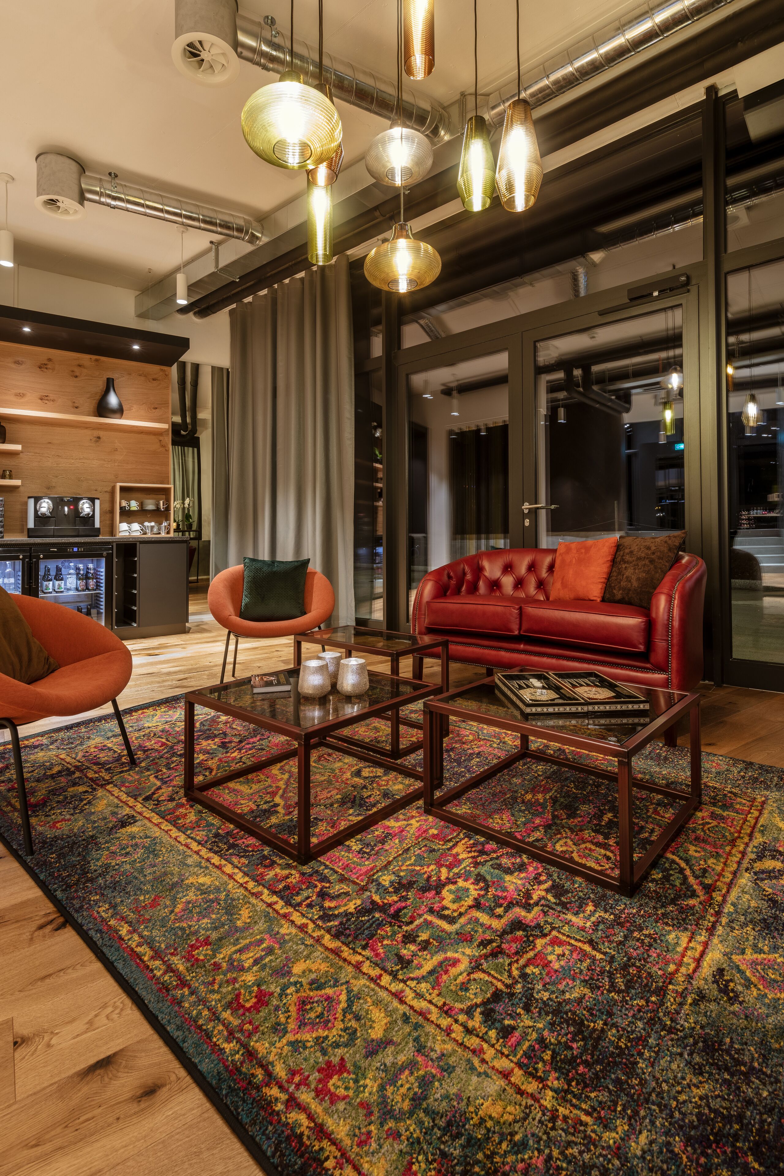 Ein modernes Wohnzimmer mit einem roten Ledersofa, einem Glastisch und einem farbenfrohen Teppich. Über dem Sofa hängen Pendelleuchten und im Hintergrund ist eine Küche zu sehen.
