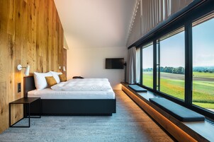 A modern bedroom with a large bed, wooden panel walls, a TV, and a view of a green landscape.