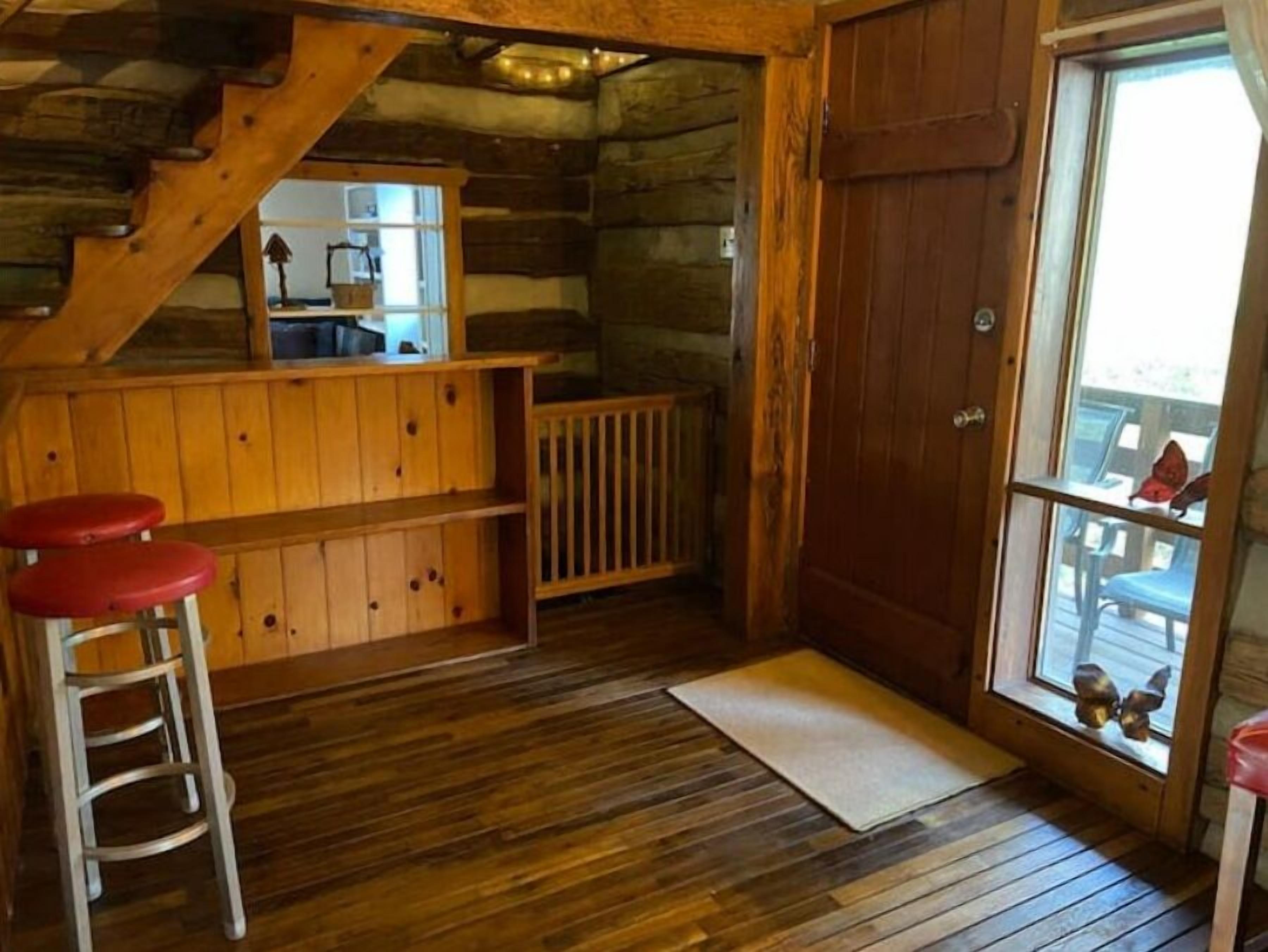 A rustic cabin interior with a wooden floor, a wooden bench, a small stool, a wooden door, and a window with a view of outdoor furniture.