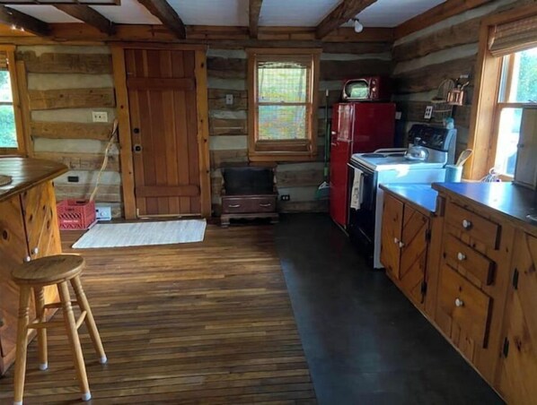 A log cabin interior with a wooden dining table, a stool, a red refrigerator, and a kitchen area with wooden cabinets.