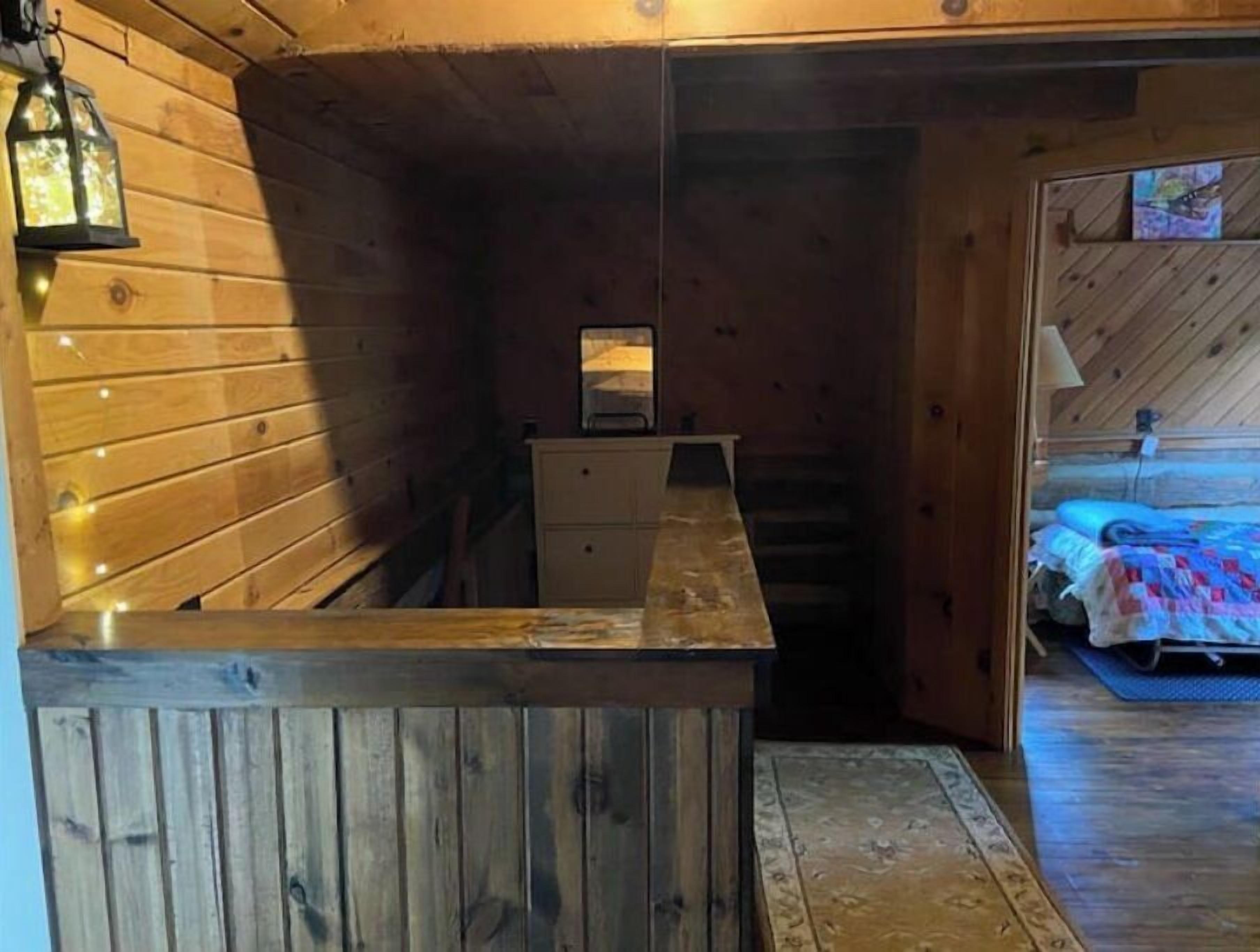 Interior de una cabaña de madera con una pequeña kitchenette, una cama con manta azul y un banco de madera.