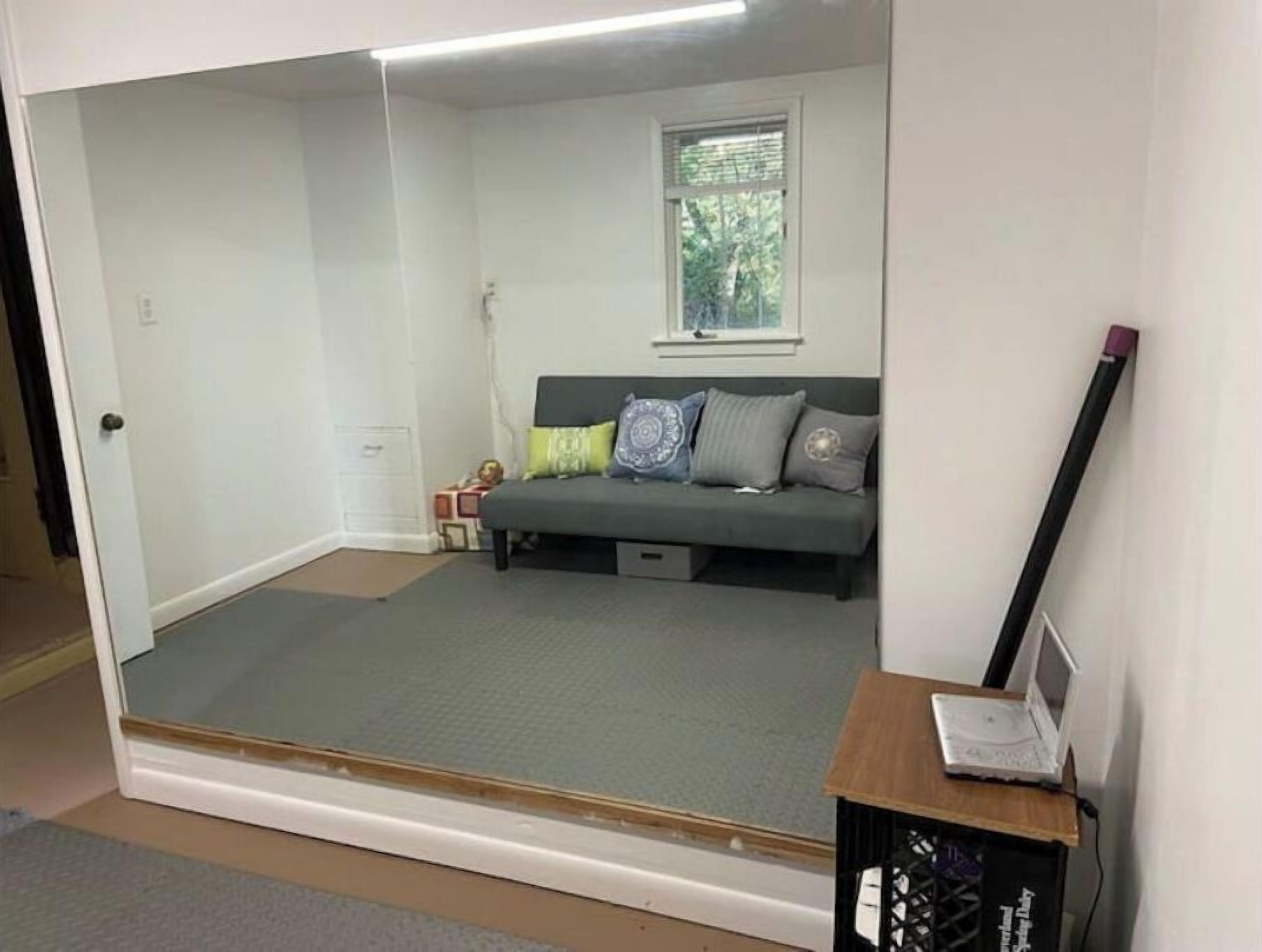 Una habitación compacta con una puerta corredera que da a un espacio más amplio, un sofá gris con cojines estampados, una pequeña mesa de madera con una computadora portátil y una ventana con persianas.
