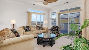 A spacious living room with a beige sofa, two armchairs, a round coffee table, and a ceiling fan. Large windows provide natural light and views of the city.