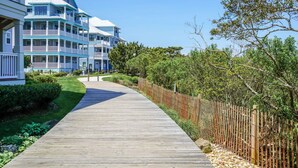 A wooden boardwalk leading to a multi-story building with balconies, surrounded by greenery and a wooden fence.