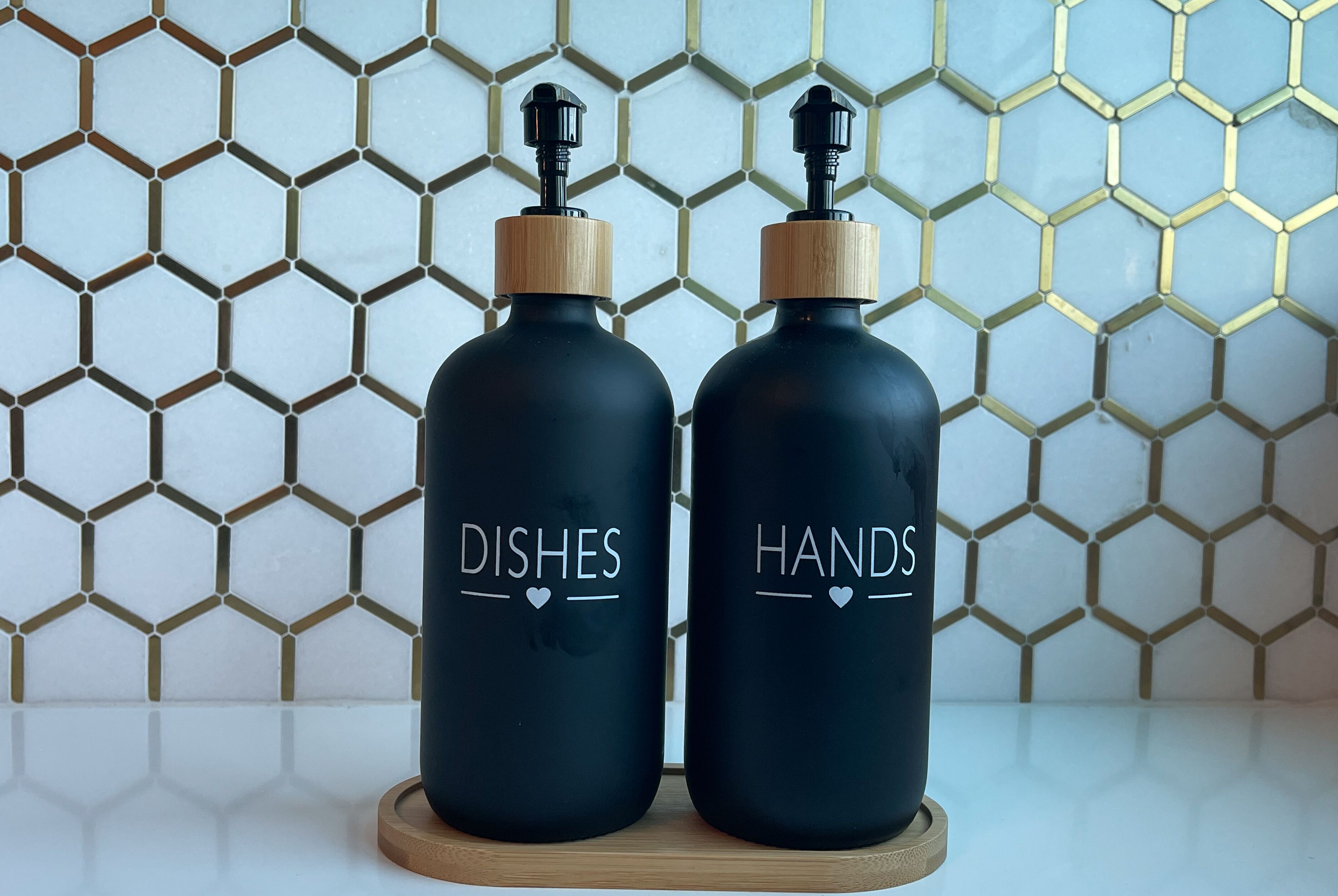Two black bottles with wooden caps labeled "DISHES" and "HANDS" on a wooden coaster against a tiled wall with a honeycomb pattern.