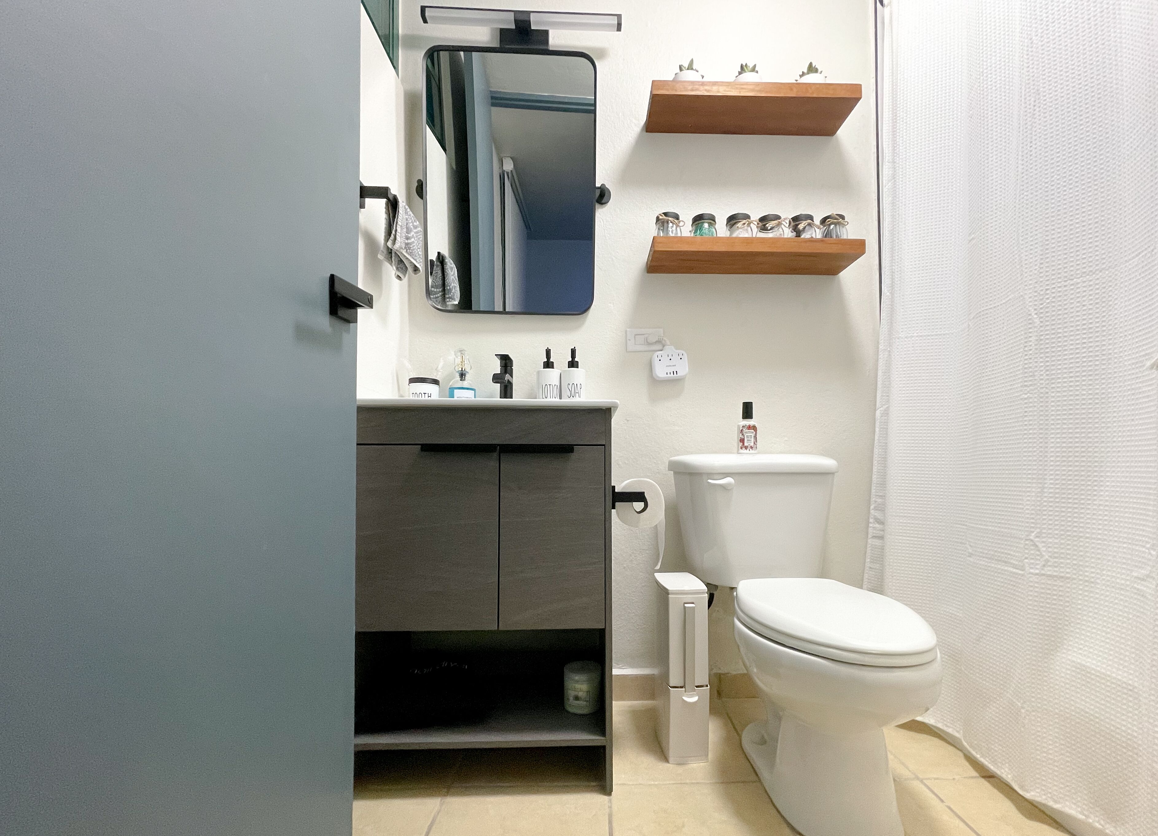 A modern bathroom with a toilet, sink, and mirror. There are wooden shelves above the sink and a towel hanging on the wall.