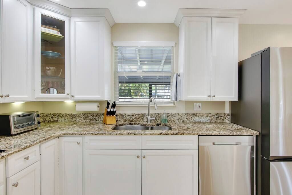 A kitchen with white cabinets, a granite countertop, a stainless steel refrigerator, and a window with blinds.