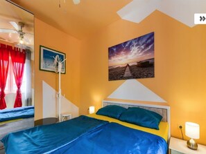 Hotelkamer met een bed, nachtkastje, lamp, schilderij van een zonsondergang, kapstok en raam met rode gordijnen.