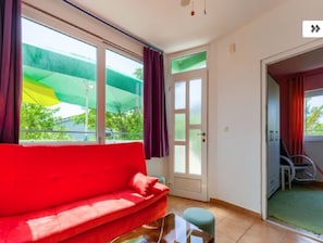 Een kamer met een rode bank, een glazen tafel, een deur met geëtste glaspanelen en uitzicht op groen buiten.