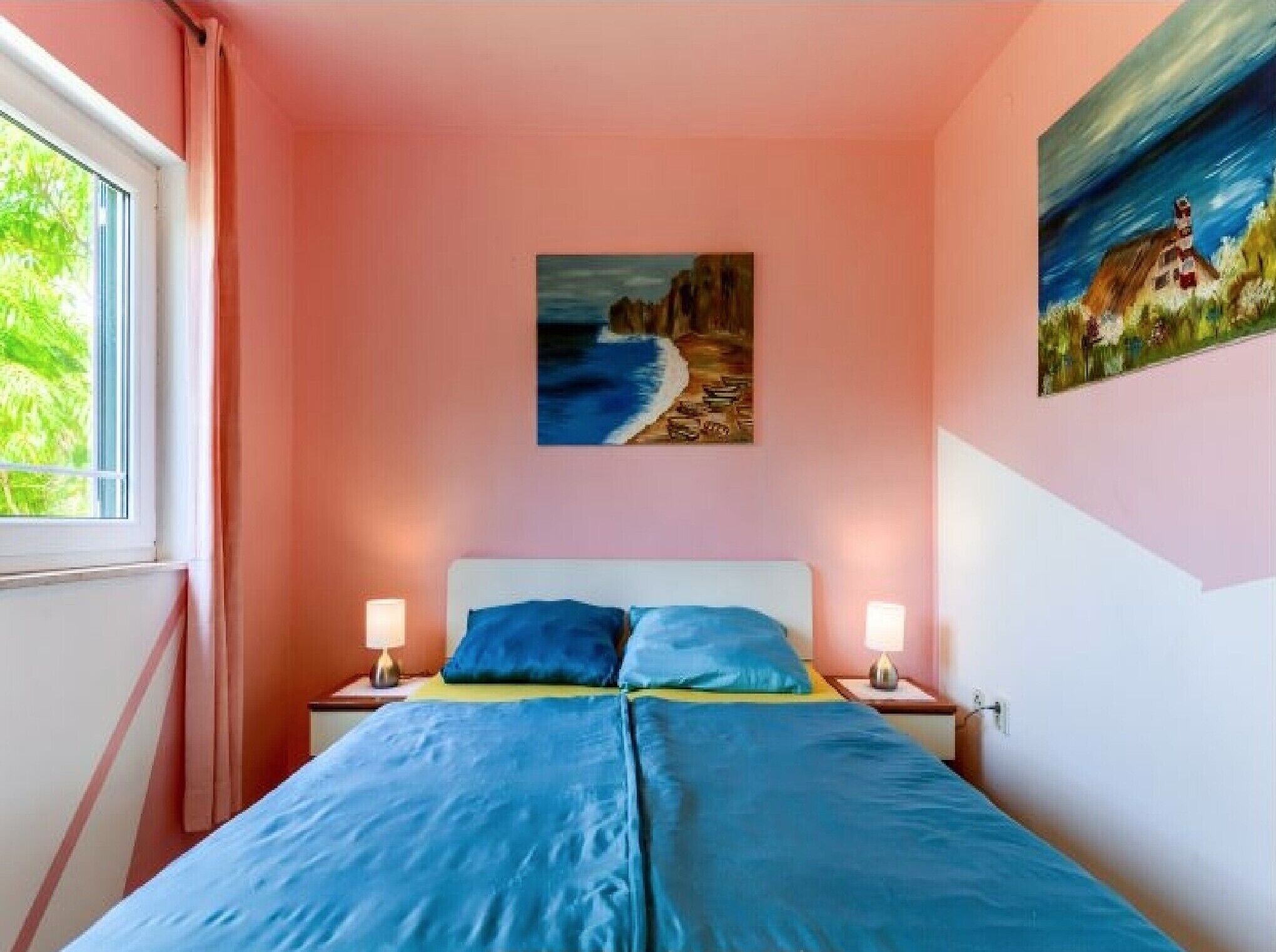 A bedroom with a bed, two bedside tables, a painting of a beach scene, and a window with curtains.