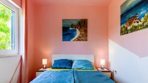 Un dormitorio con una cama, dos mesitas de noche, un cuadro de una playa y una ventana con cortinas.