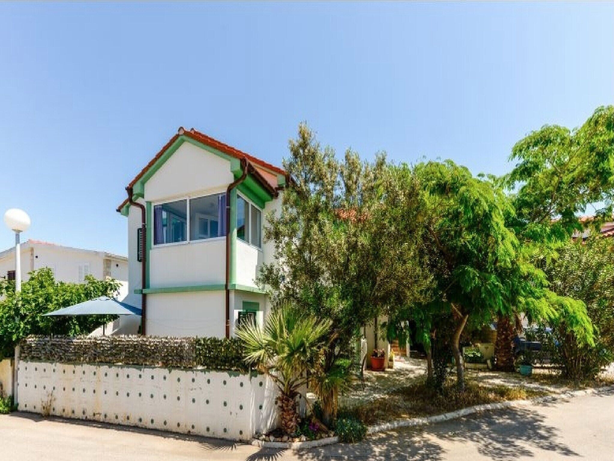 A two-story building with a white facade and green trim, surrounded by lush greenery and a paved area.