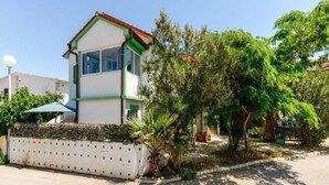 Un edificio de dos plantas con fachada blanca y detalles verdes, rodeado de abundante vegetación y una zona pavimentada.