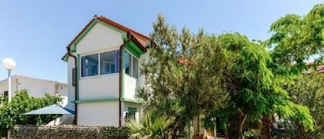 A two-story building with a white facade and green trim, surrounded by lush greenery and a paved area.