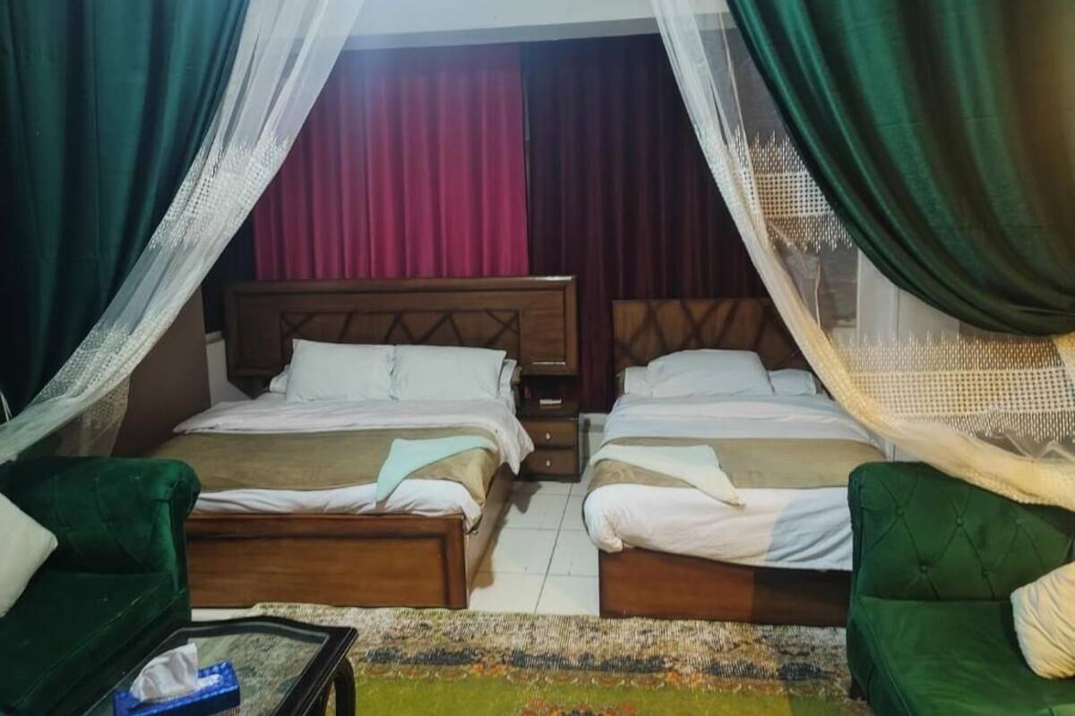 A hotel room with two beds, a green sofa, a bedside table, and a window with curtains.