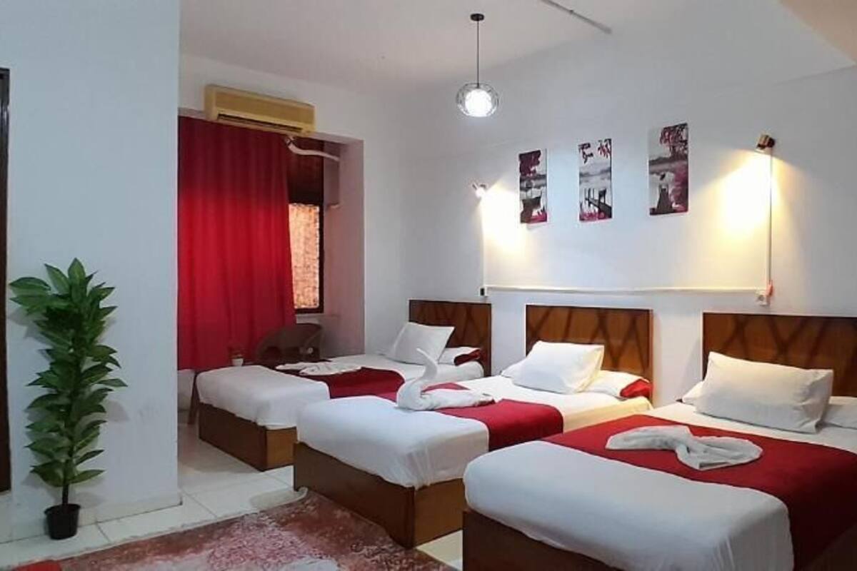 A hotel room with three beds, a potted plant, a red curtain, and wall art.