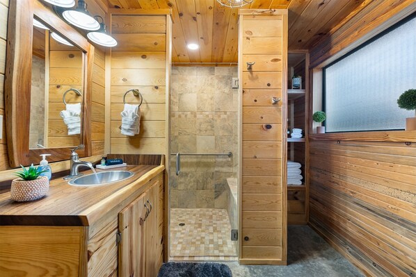 A wooden cabin-style bathroom with a shower, a sink with a mirror, and a towel rack.