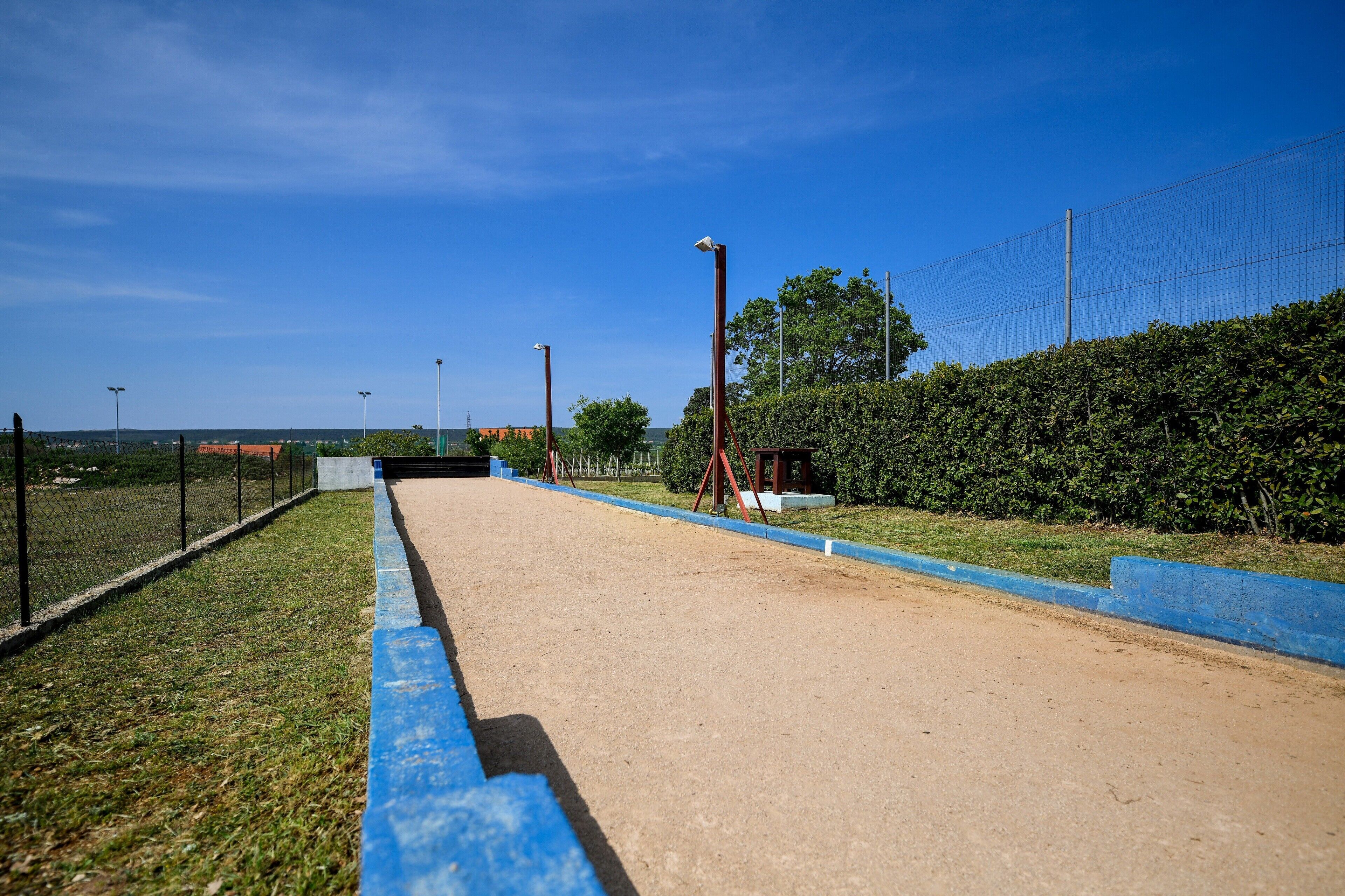 A clear, sunny day at a sports facility with a dirt track, blue safety barriers, and a chain-link fence.