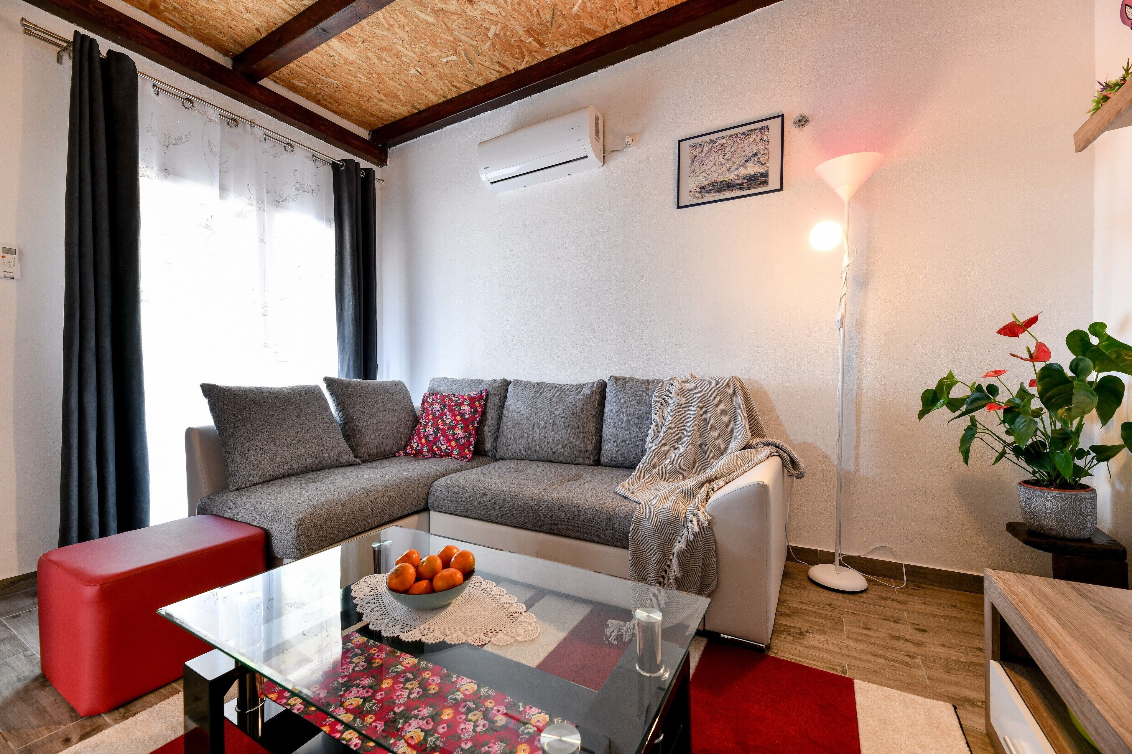 Een moderne woonkamer met een grijze hoekbank, een glazen salontafel met bloemenonderzetter, een rode poef en een kamerplant.