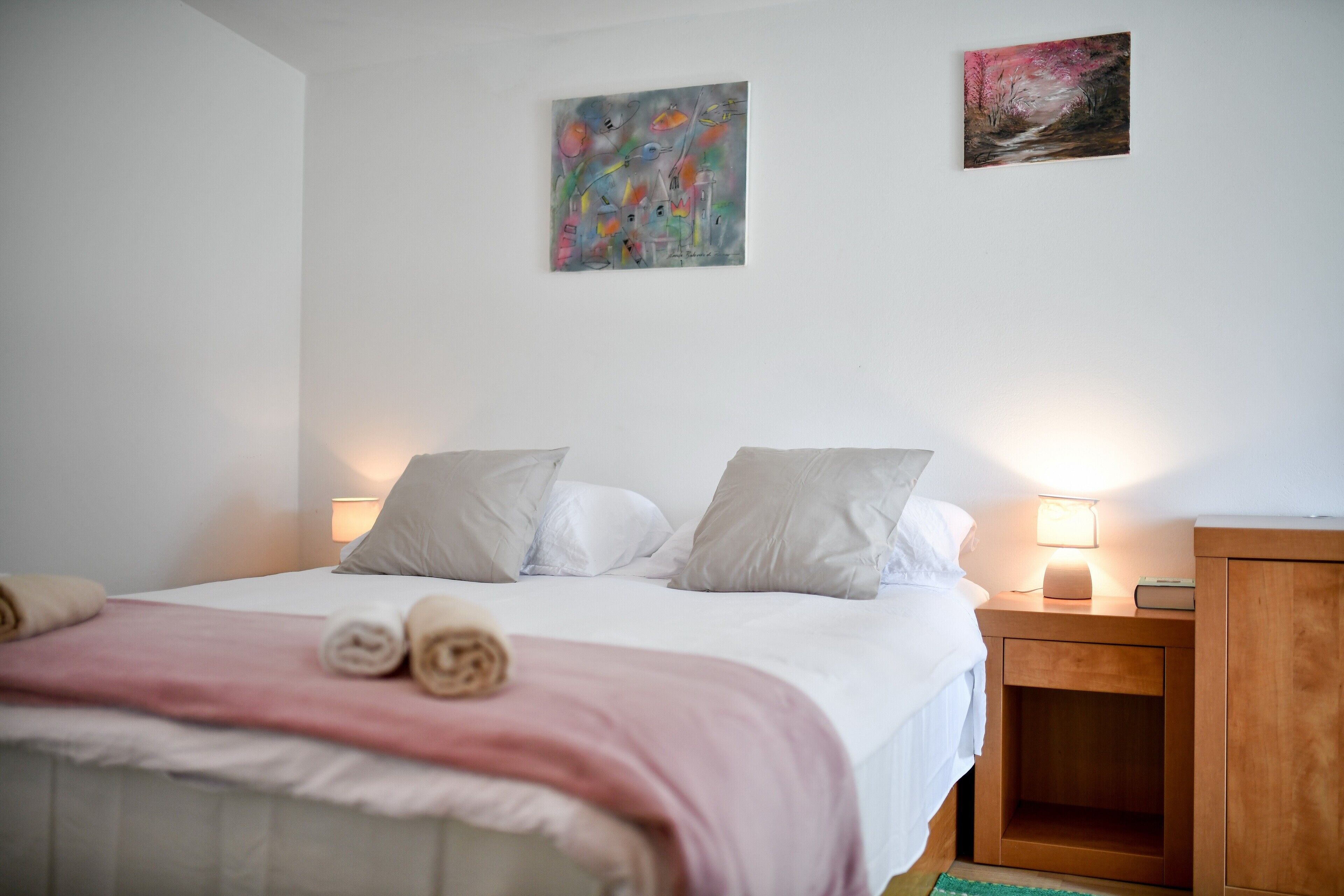 En snyggt bäddad säng med ett rosa överkast och hoprullade handdukar, två sänglampor och ett träbord vid sängen med en lampa.