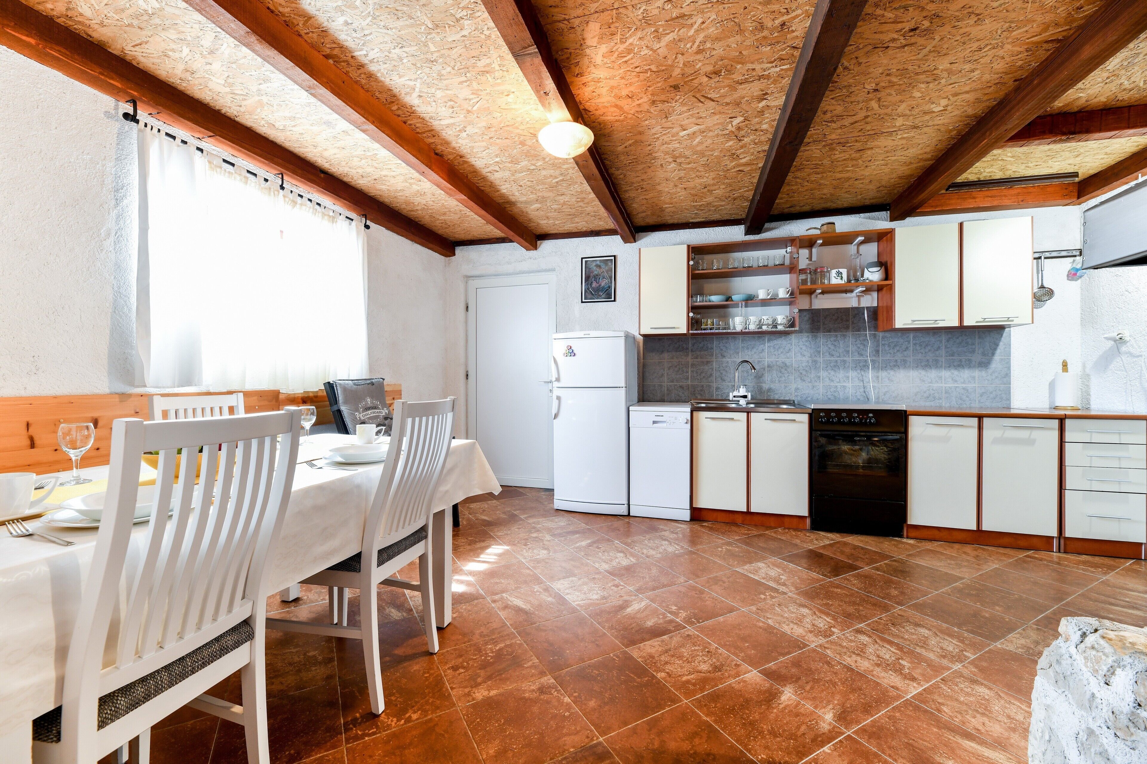 A kitchen with white cabinets, a white refrigerator, and a dishwasher. There is a dining table with white chairs and a window with curtains.