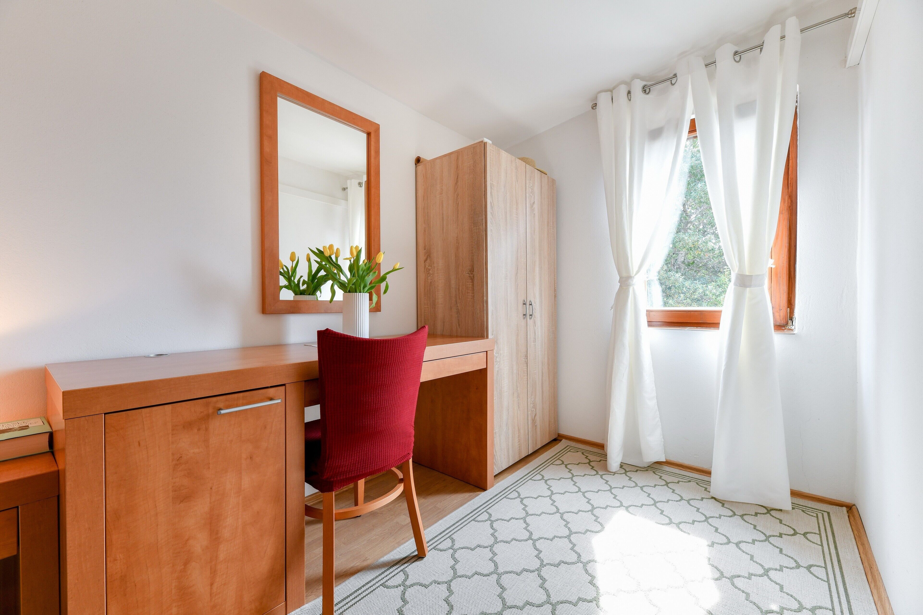 Ein Zimmer mit einem Holztisch, einem roten Sessel, einem Spiegel, einem Kleiderschrank und einem Fenster mit weißen Vorhängen.