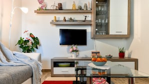 Una sala de estar moderna con un sofá, una mesa de centro de vidrio, un televisor, un mueble bajo de madera y objetos decorativos en estanterías.